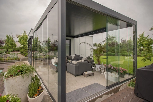 Bioklimatická pergola Renson od PLAS.cz s prosklenými stěnami a moderním venkovním posezením, která majitele chrání před nepříznivým počasím.