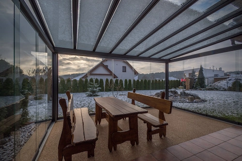 Nezateplená prosklená pergola od PLAS.cz s dřevěným posezením, nabízející útulné venkovní stolování i v zimním období.