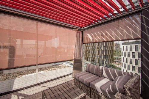 Bioklimatická pergola Renson od PLAS.cz s červenými lamelami a prosklenými stěnami, která vytváří elegantní venkovní posezení na terase s výhledem na město.